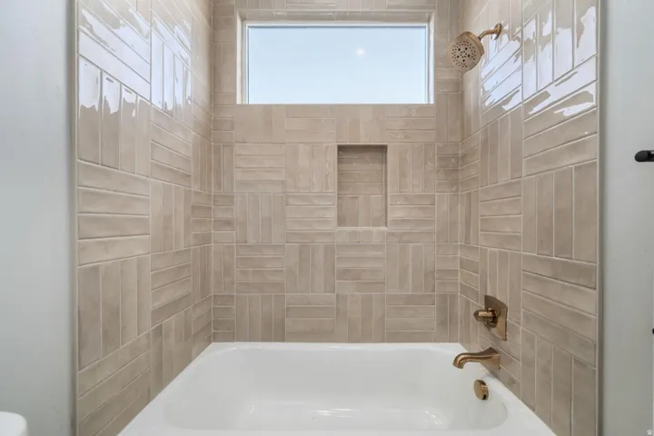 Subway tile surround in a natural tone, built-in recessed shelf, white bathtub, bronze-finish fixtures, and transom window
