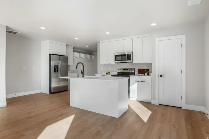 White shaker cabinetry, stainless steel appliances, central island with sink, wood-finish flooring, and recessed lighting