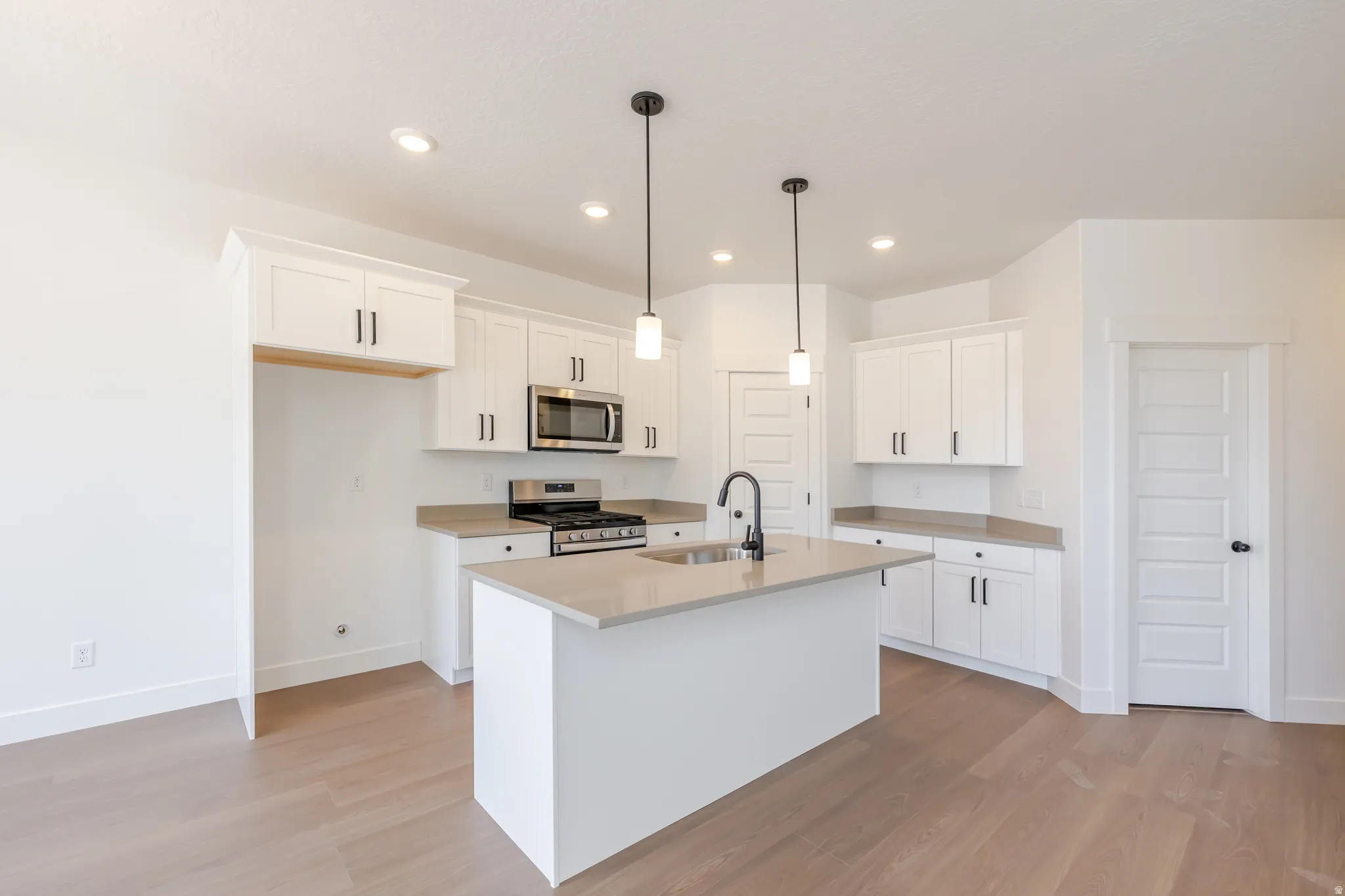 Modern kitchen featuring light wood-finish flooring, white cabinetry with dark hardware, a central island with an undermount sink, pendant lighting, and stainless steel appliances