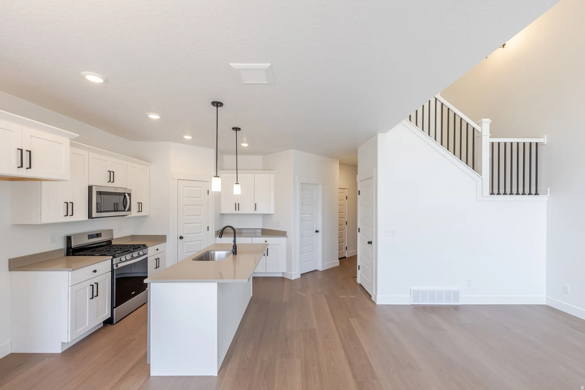 Open-concept kitchen featuring an island with a sink and pendant lighting, white cabinetry with black hardware, stainless steel appliances, and light-toned countertops