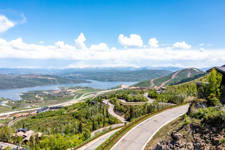 Expansive mountain and lake landscape featuring a winding paved road, distant snow-capped peaks, and scattered residential properties