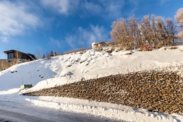 Hillside property featuring a modern architectural design with large windows, a gabion wall retaining structure, and a snow-covered landscape