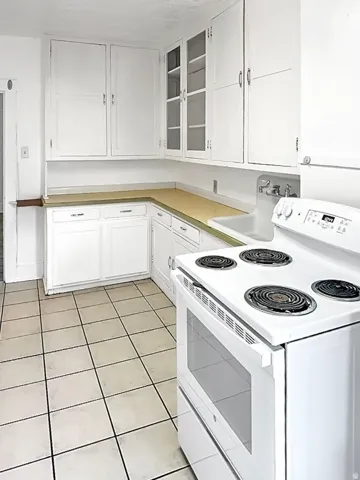 Kitchen with white range with electric stovetop, white cabinets, glass insert cabinets, light tile patterned floors, and light countertops