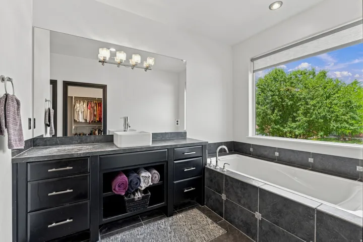 Bathroom featuring a built-in soaking tub with dark tile surround, a large window, and a vanity with a vessel sink