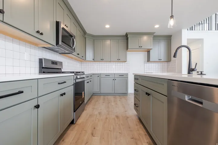 Kitchen featuring light green cabinetry with black hardware, white subway tile backsplash, light-colored countertops, stainless steel appliances, and wood-finish flooring