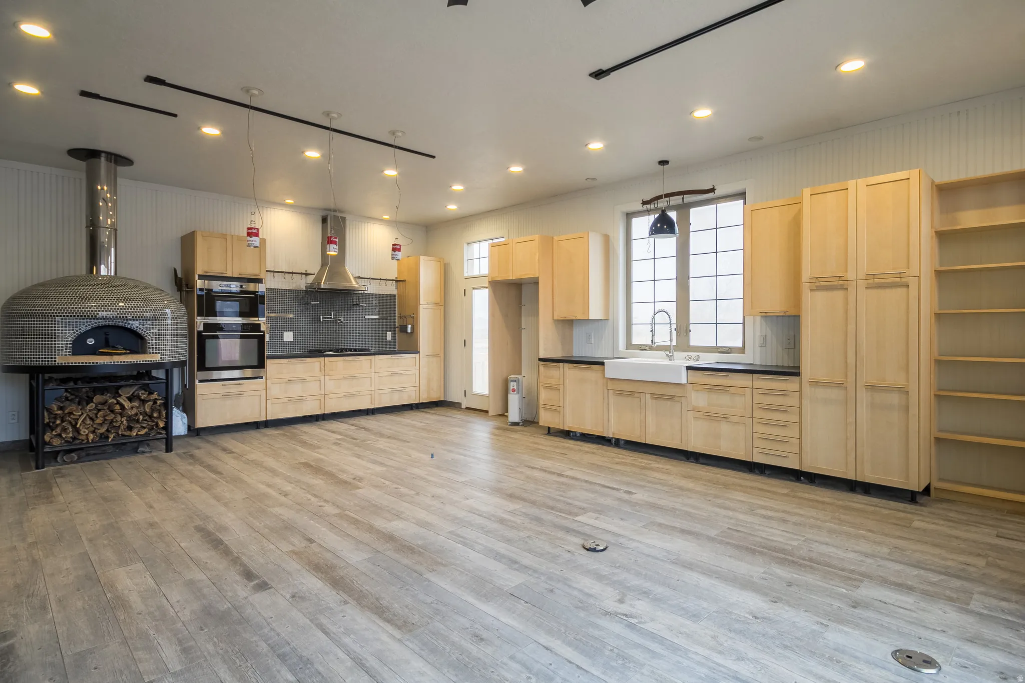 Kitchen with light wood finish cabinetry, dark countertops, open shelves, hanging light fixtures, and double oven