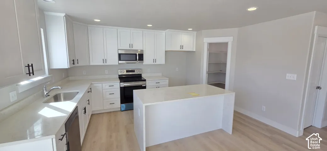 White cabinetry with matte black hardware, stainless steel appliances, light-toned countertops, a central island, and wood-finish flooring