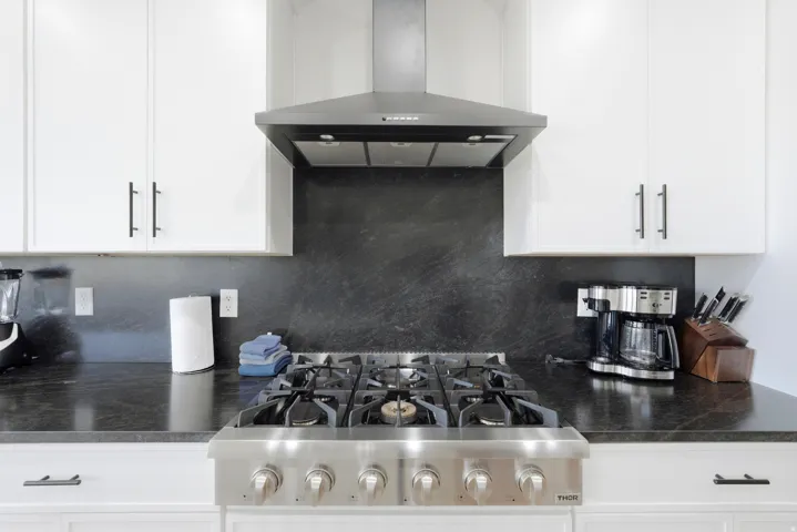 Stainless steel range with a built-in exhaust hood, white upper cabinetry with black hardware, and dark countertops with a coordinating backsplash