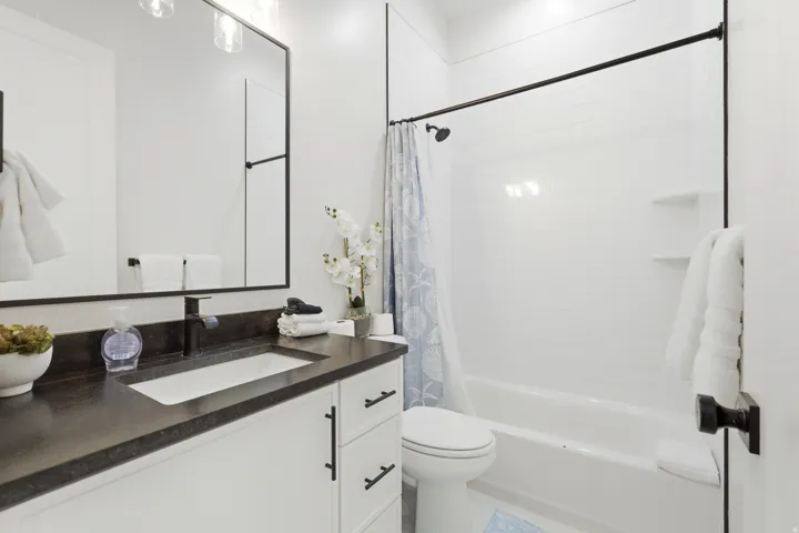 Vanity with dark countertop and undermount sink, white cabinetry with dark hardware, framed mirror, and a full bathtub with shower