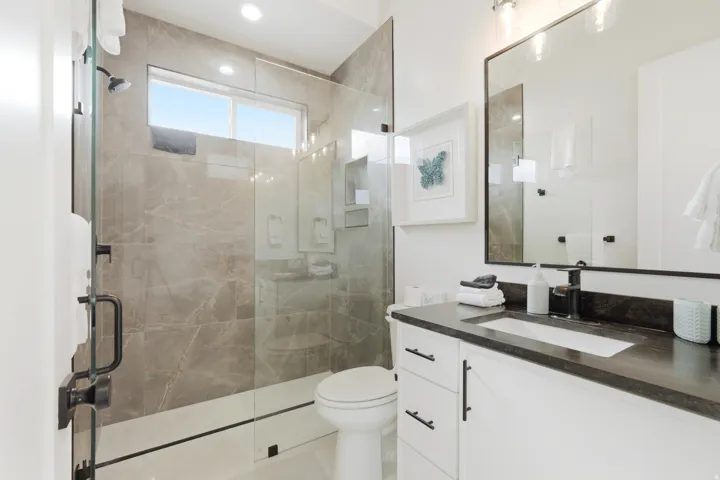 Frameless glass shower enclosure with gray veined tile surround, white vanity with dark countertop, rectangular undermount sink, and black fixtures