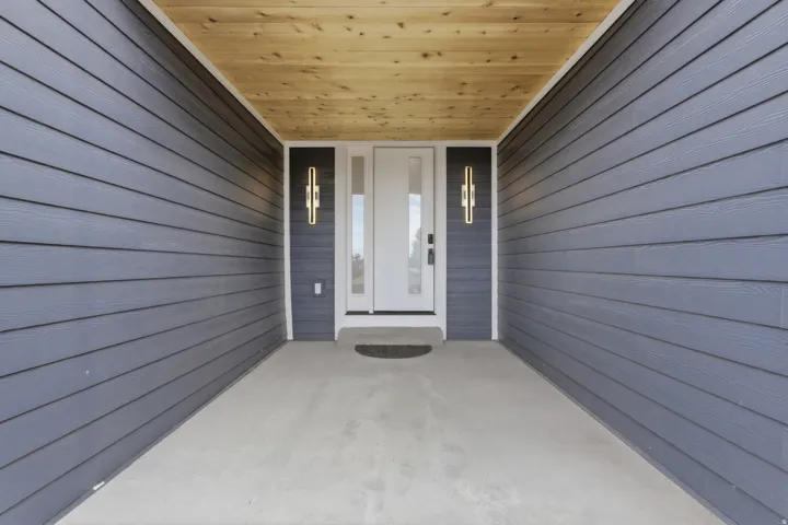 Covered entryway featuring light wood-finish ceiling, grey horizontal siding, and modern wall sconces flanking the entry door