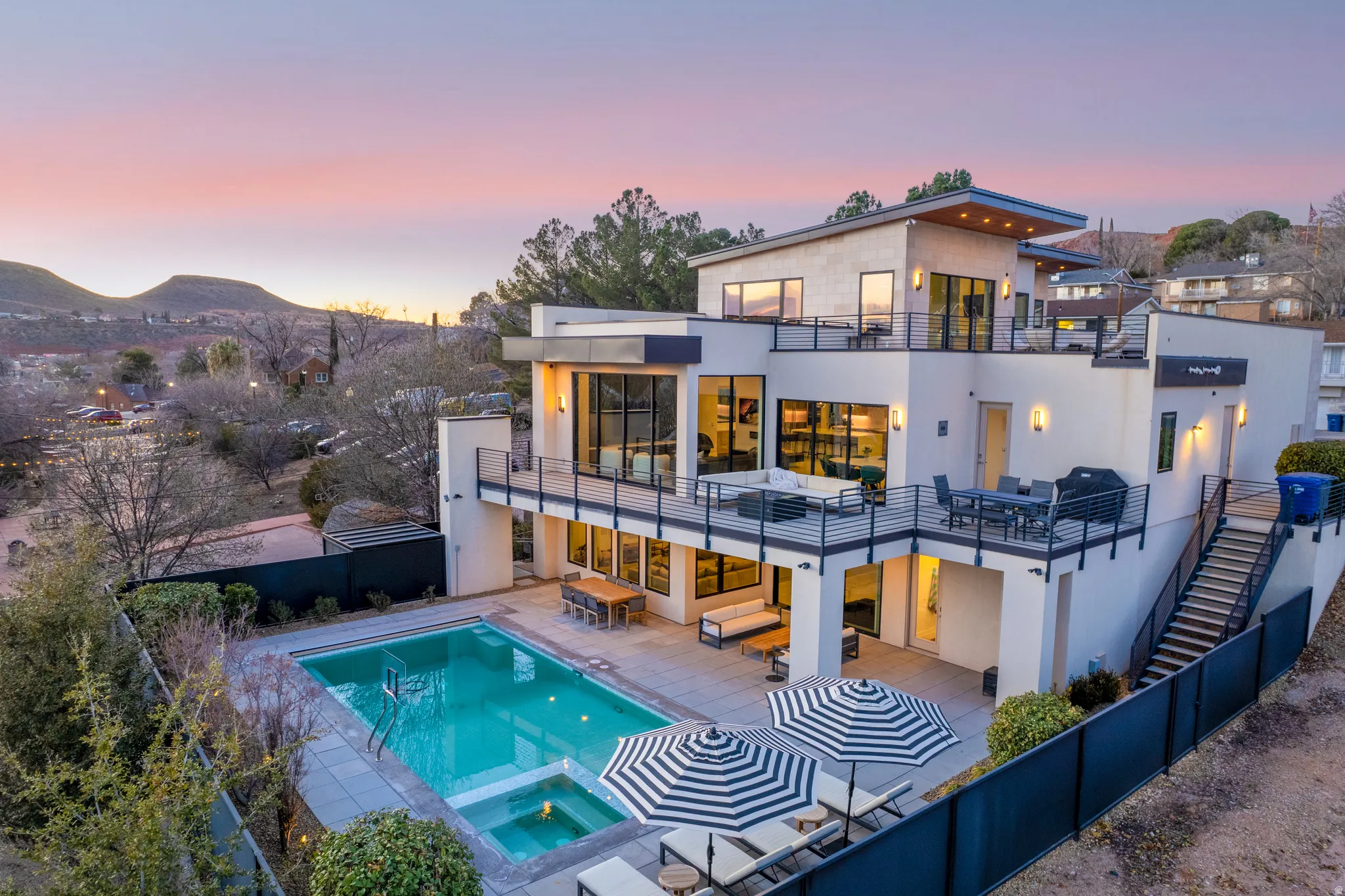 Back of house at dusk featuring stucco siding, a pool with connected hot tub, a balcony, a fenced backyard, and a patio area