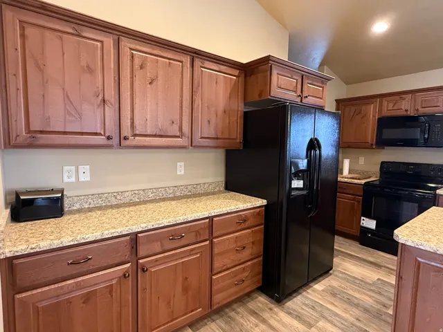 Kitchen featuring black appliances, light wood-style flooring, vaulted ceiling, light stone counters, and wood finish cabinets