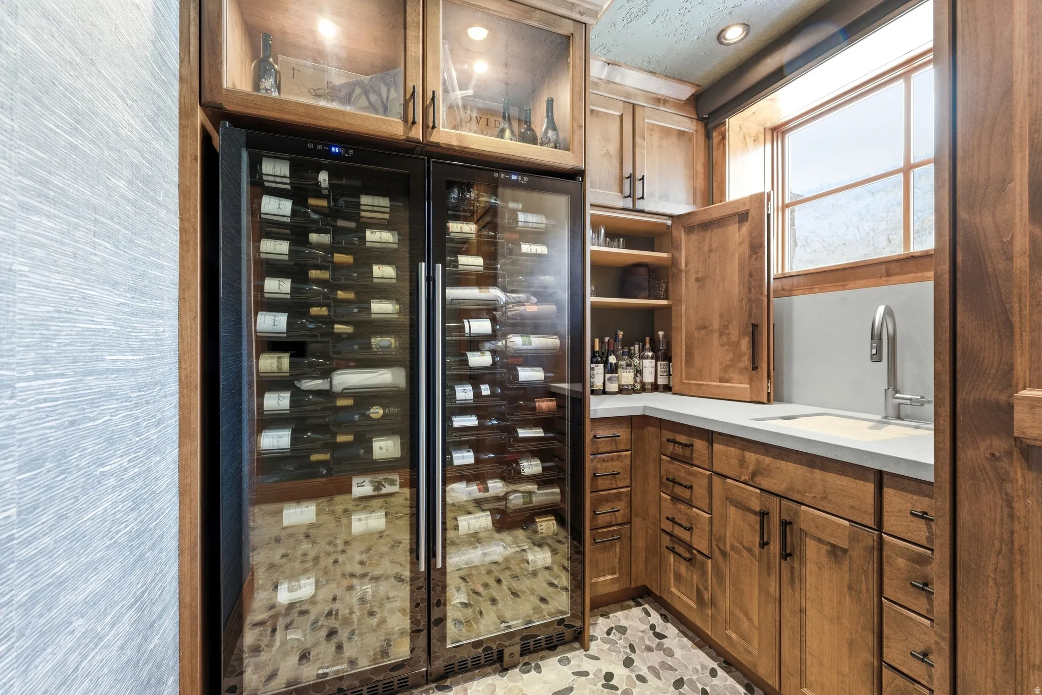 Built-in wine storage with dual-zone refrigerators, custom wood cabinetry, stone countertop with integrated sink, and an elegant stone pebble floor