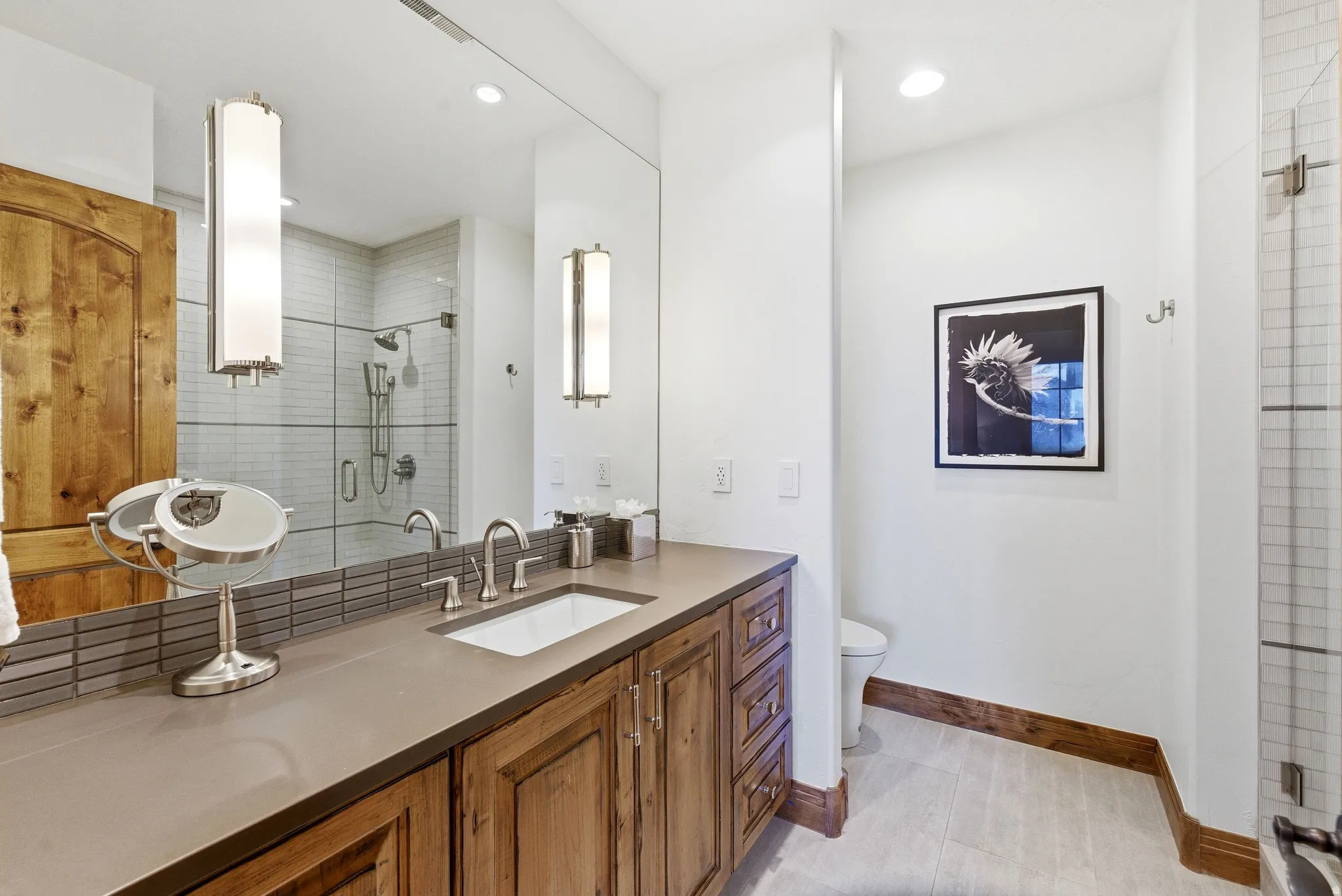 Spacious bathroom featuring a wood-finish vanity with an integrated sink, a large wall-mounted mirror, a glass-enclosed shower with white subway tile, and light-toned wood-finish flooring