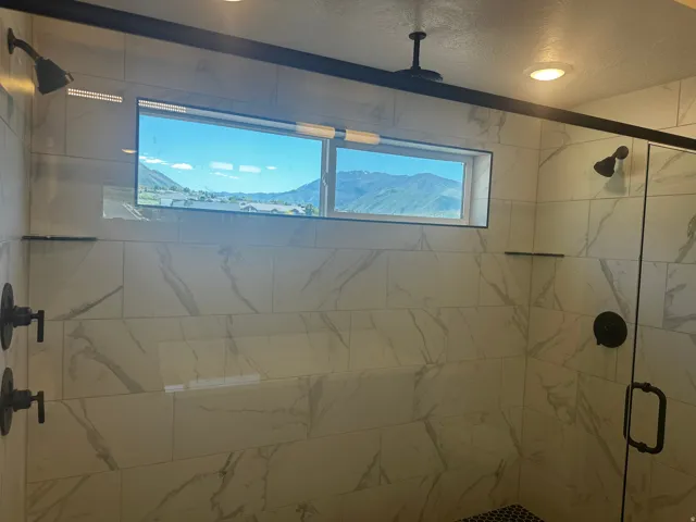 Shower enclosure featuring large format white and gray marbled tile, dark bronze fixtures, glass shower door, and a built-in window
