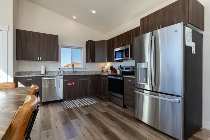 Kitchen with dark wood finish cabinets, stainless steel appliances, vaulted ceiling, dark wood-style floors, and light stone counters