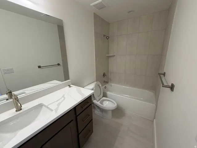 Bathroom featuring a dark wood-finish vanity with a white countertop and integrated sink, a full-width mirror, a bathtub with a tiled shower surround, and light-toned floor tiling