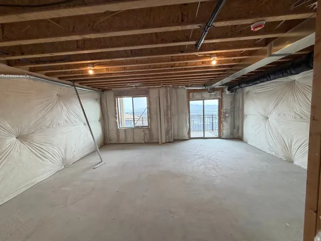 Unfinished room featuring exposed wooden ceiling joists, two windows, and a sliding glass door with a metal railing
