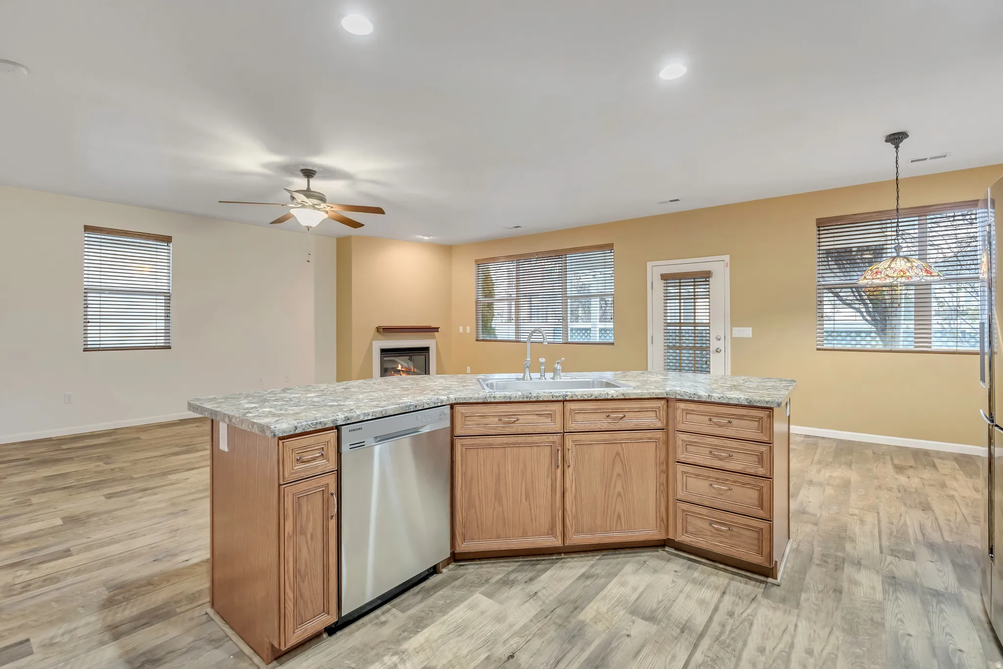 Kitchen featuring a center island with sink, a warm lit fireplace, dishwasher, wood finish cabinets, and pendant lighting