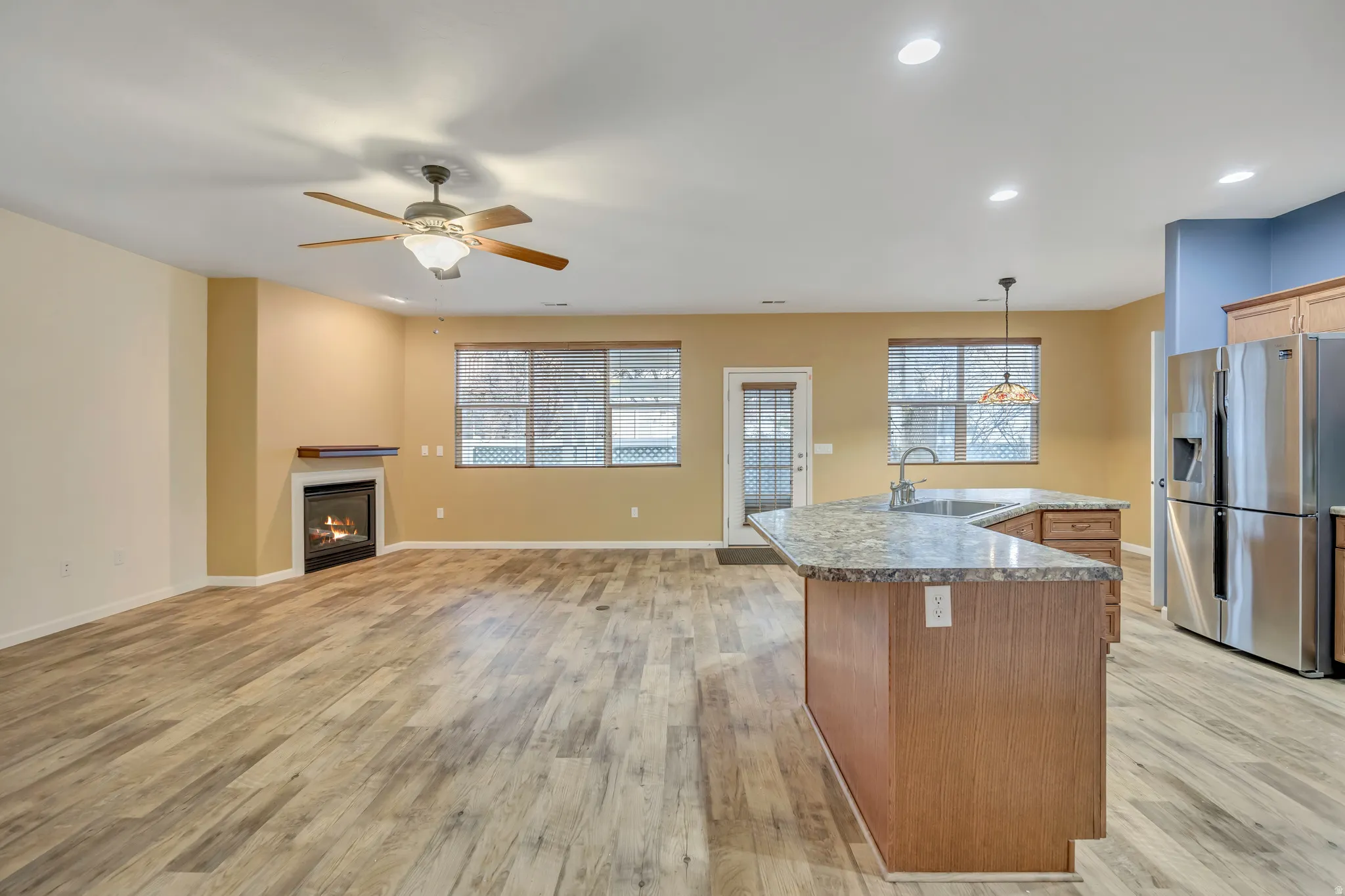 Kitchen featuring stainless steel fridge with ice dispenser, hanging light fixtures, a glass covered fireplace, an island with sink, and light wood-type flooring