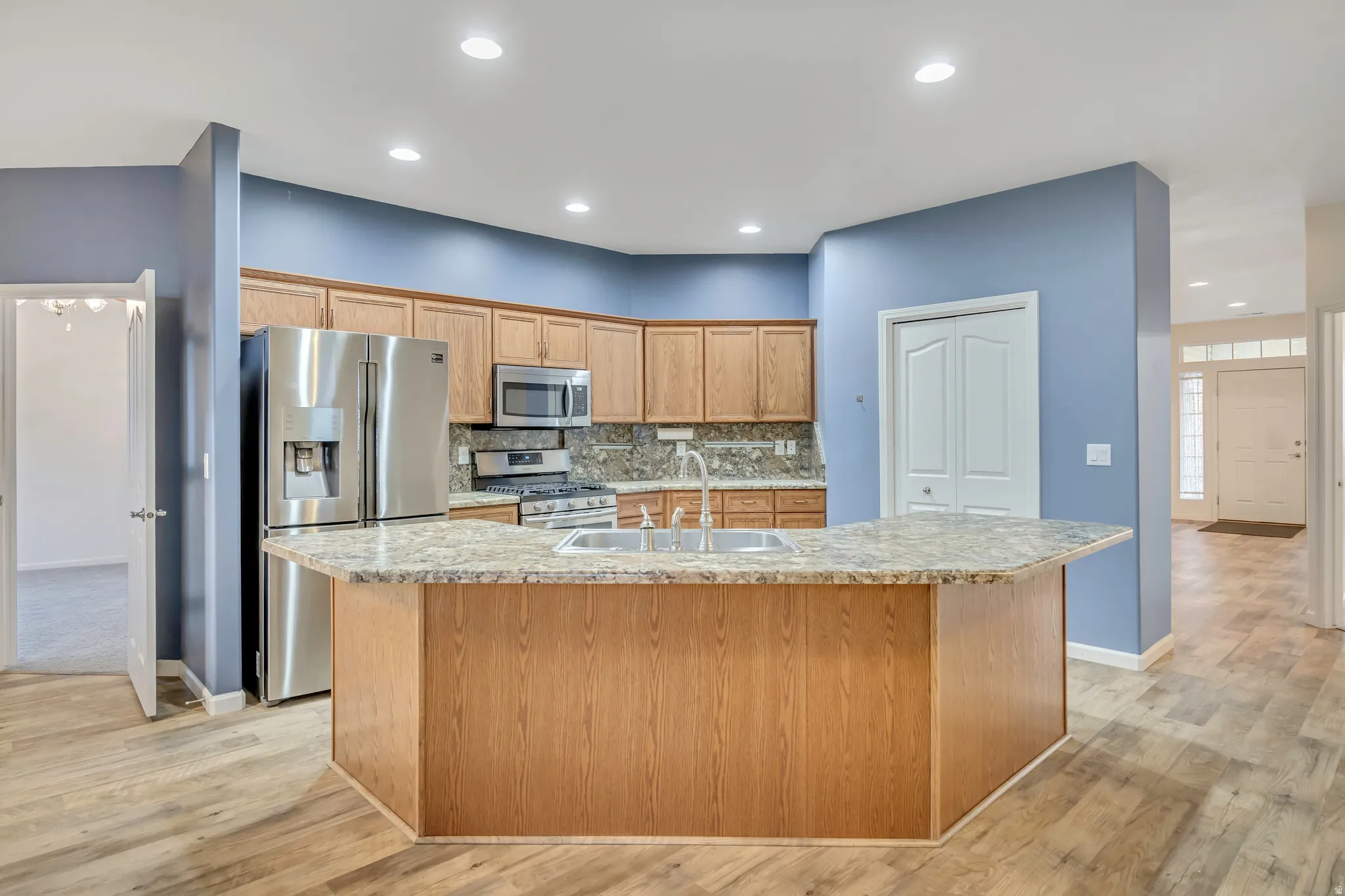 Kitchen with stainless steel appliances, an island with sink, decorative backsplash, light stone countertops, and light wood finished floors