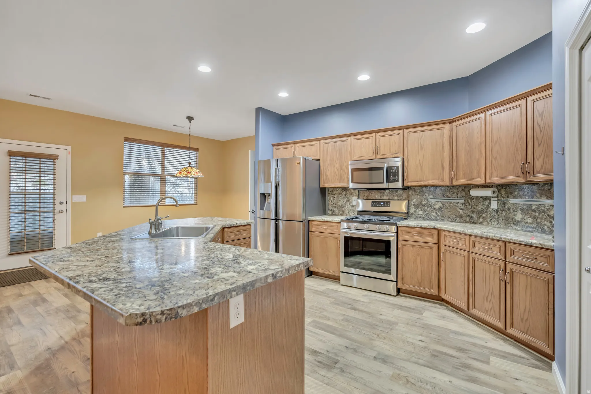 Kitchen with stainless steel appliances, an island with sink, hanging light fixtures, decorative backsplash, and light wood-type flooring