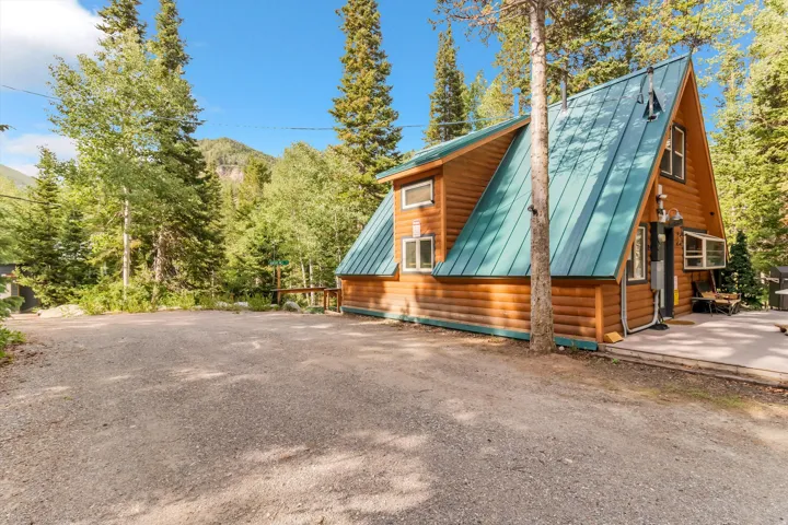 A-frame wood cabin featuring a teal metal roof, log siding, a front patio, and a gravel driveway