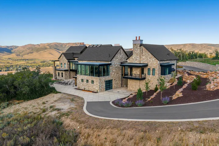 Stone exterior with dark shingle roofing, expansive glass-enclosed sections, and a paved driveway leading to the entrance