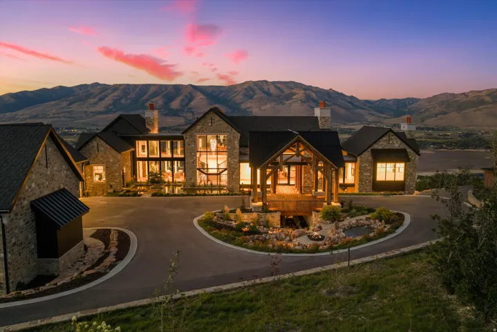 Stone and dark-pitched roof exterior with extensive architectural lighting, a circular driveway, an elevated timber-framed entry, and multiple chimneys
