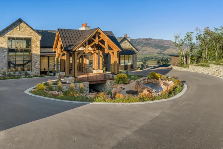 Stone exterior with a prominent timber-frame entry, expansive windows, a circular driveway, and a decorative water feature