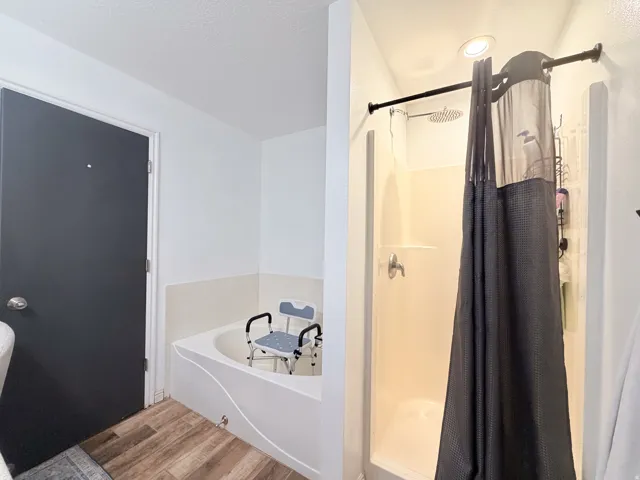 Soaking tub with white surround, separate shower with a white enclosure, overhead lighting, and wood-finish flooring
