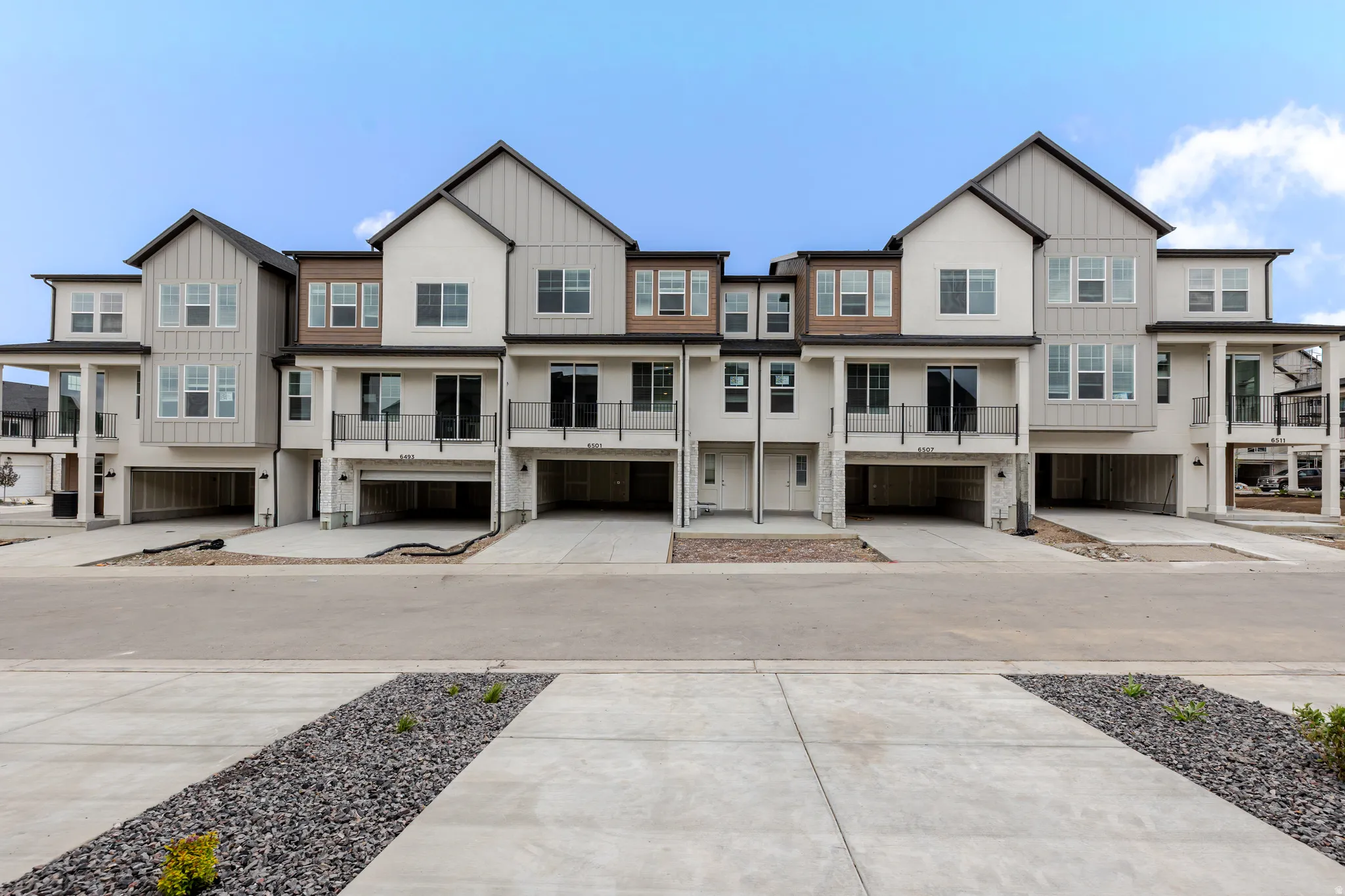Contemporary multi-level townhomes featuring stucco and vertical siding exteriors, individual garages with concrete driveways, and private balconies with black railings