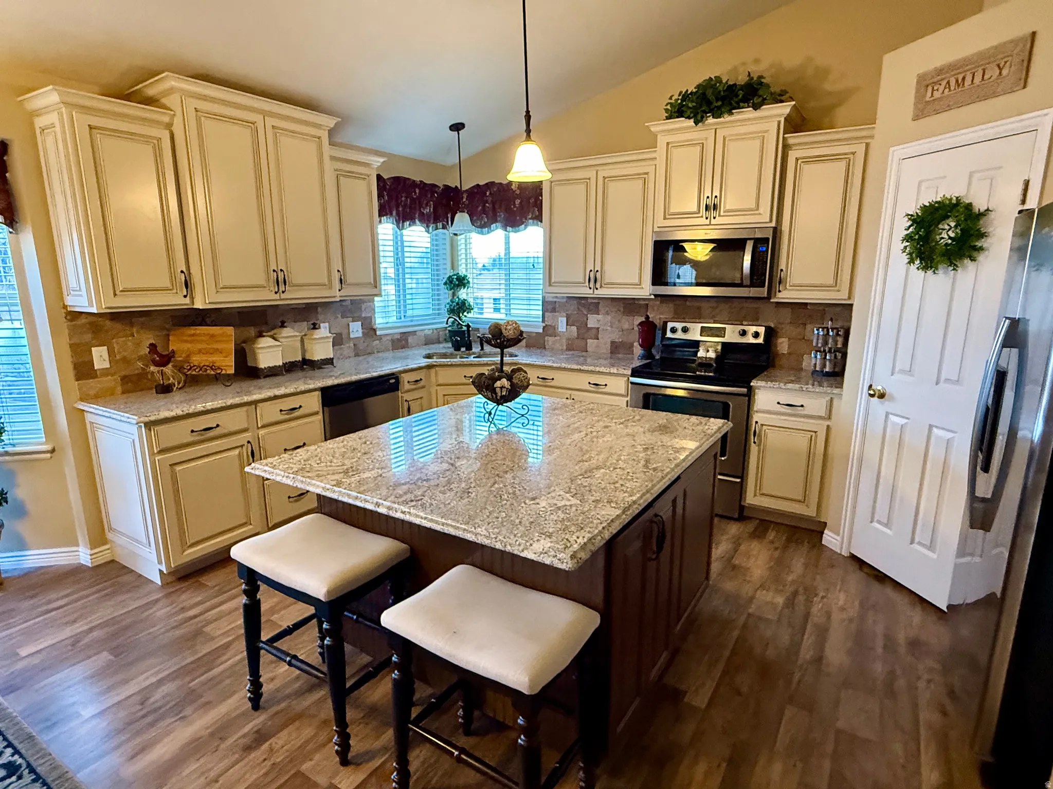 Kitchen with stainless steel appliances, cream cabinets, decorative light fixtures, lofted ceiling, and light stone countertops