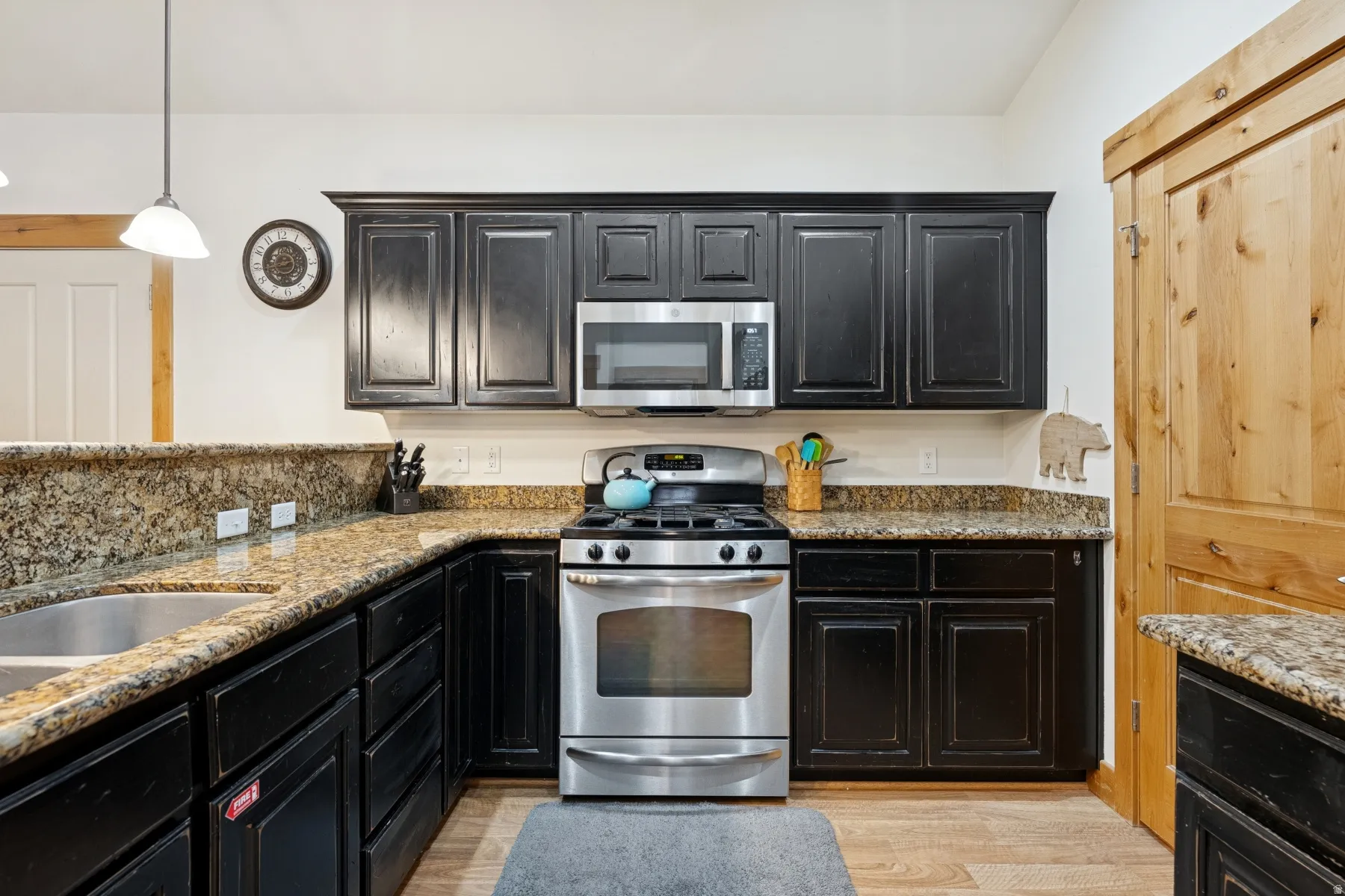 Kitchen featuring dark cabinets, stainless steel appliances, light stone counters, and light wood-style flooring