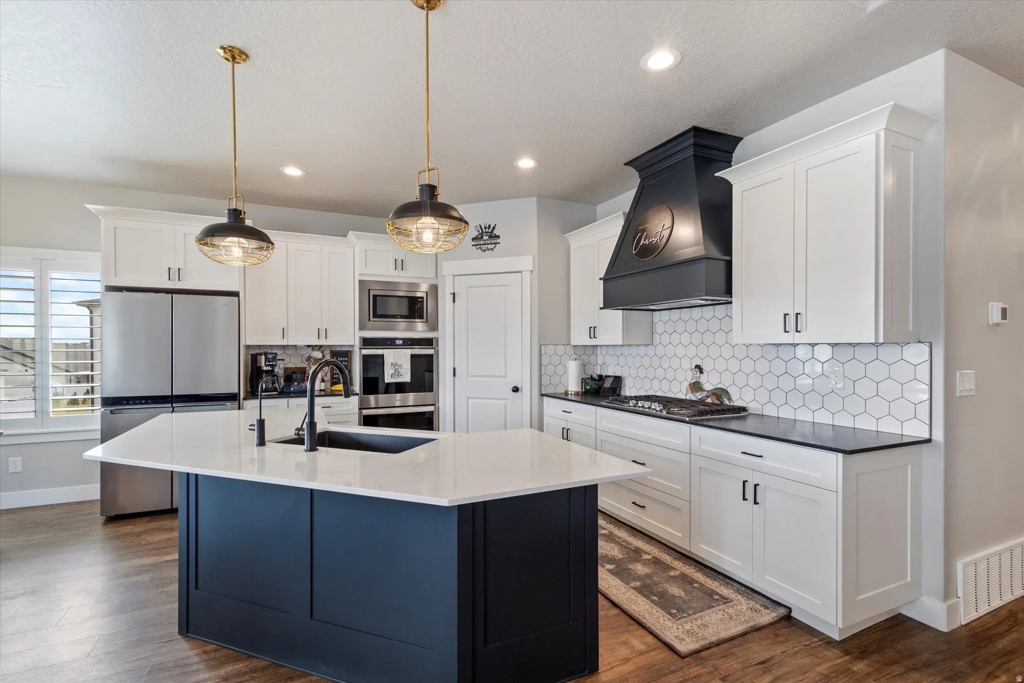 Kitchen featuring stainless steel appliances, decorative light fixtures, two tone cabinetry, an island with sink, and dark stone countertops
