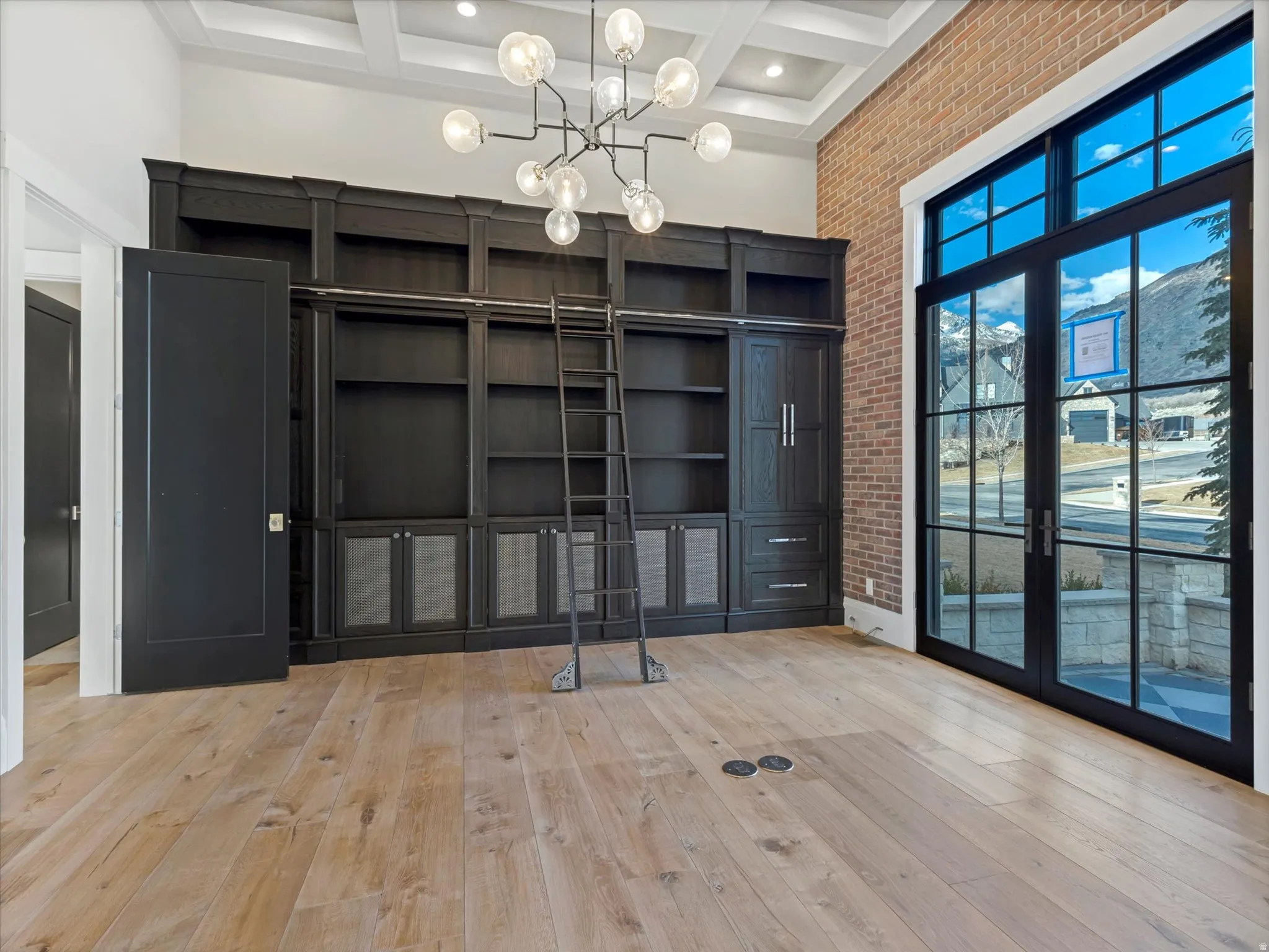 Main floor office with hidden room closed featuring coffered ceiling, suspended lighting, light wood-style floors, and brick wall