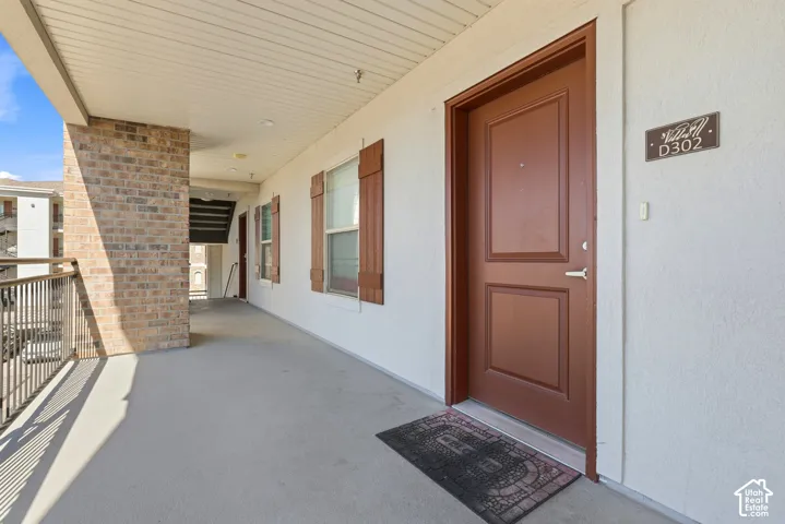 Covered exterior walkway featuring textured stucco walls, brick-clad pillar, decorative window shutters, and metal railing