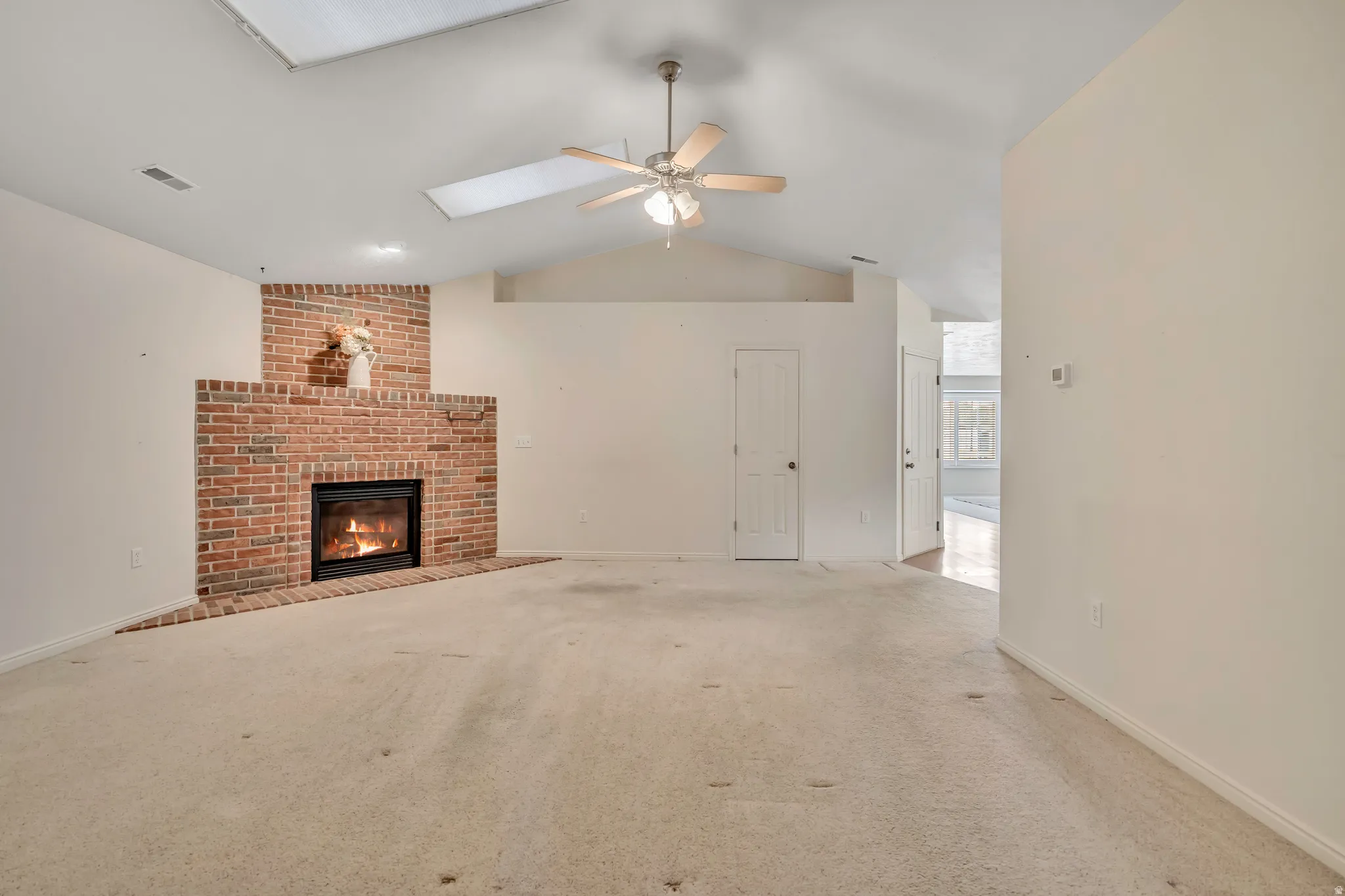 Unfurnished living room with carpet floors, a ceiling fan, lofted ceiling, a brick fireplace, and a skylight