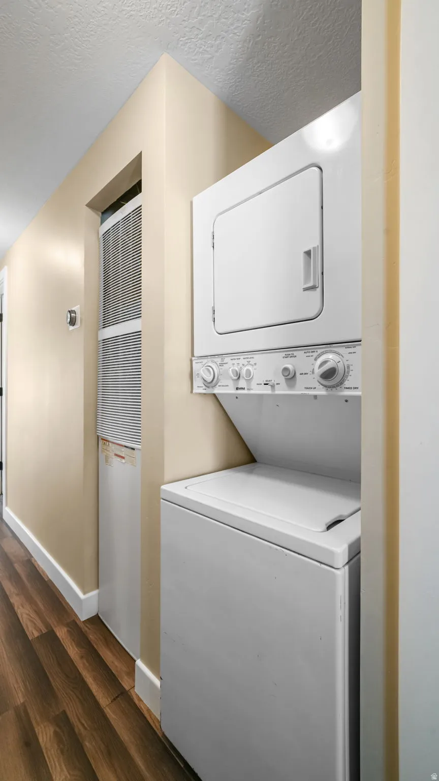 Laundry area with dark wood-type flooring, stacked washer and clothes dryer, a heating unit, and a textured ceiling