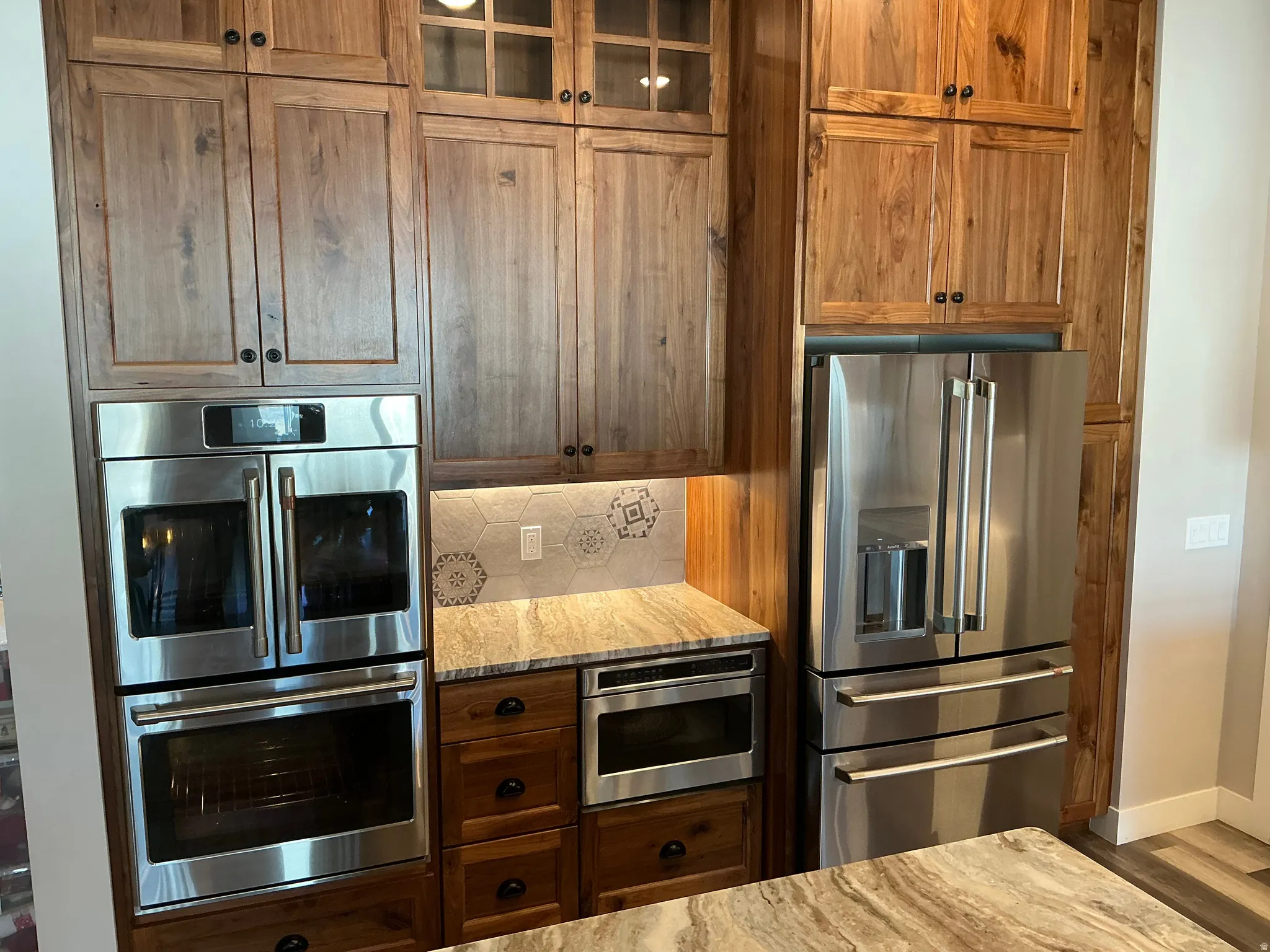 Kitchen featuring stainless steel appliances, light stone counters, backsplash, glass insert cabinets, and wood finish cabinetry