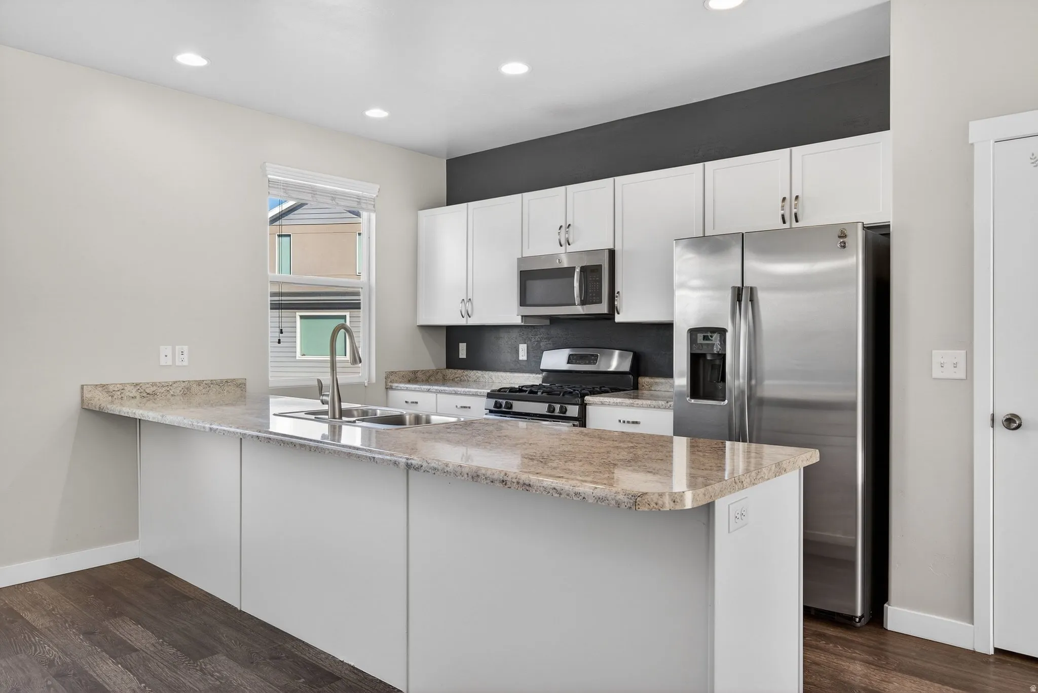 Kitchen with stainless steel appliances, a peninsula, white cabinets, dark wood-type flooring, and light stone countertops