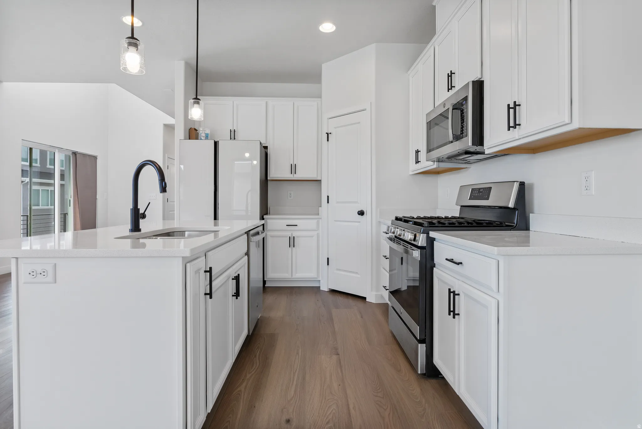 Kitchen with stainless steel appliances, dark wood finished floors, decorative light fixtures, a center island with sink, and light stone countertops