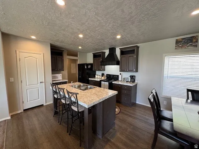 Kitchen with light countertops, a center island with sink, black appliances, recessed lighting, and dark wood finish cabinetry