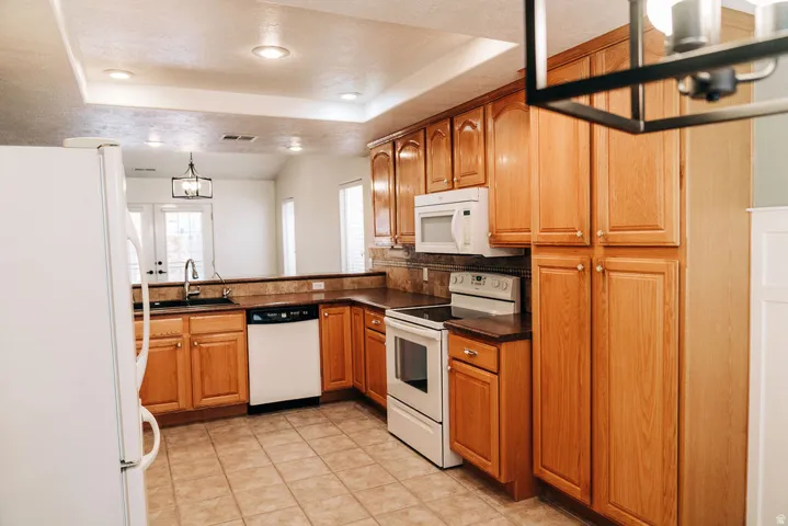 Kitchen with white appliances, dark countertops, a tray ceiling, wood finish cabinets, and a chandelier
