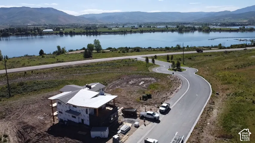 Expansive land parcel featuring an unfinished residential structure, a paved winding road, and a substantial body of water with distant mountains