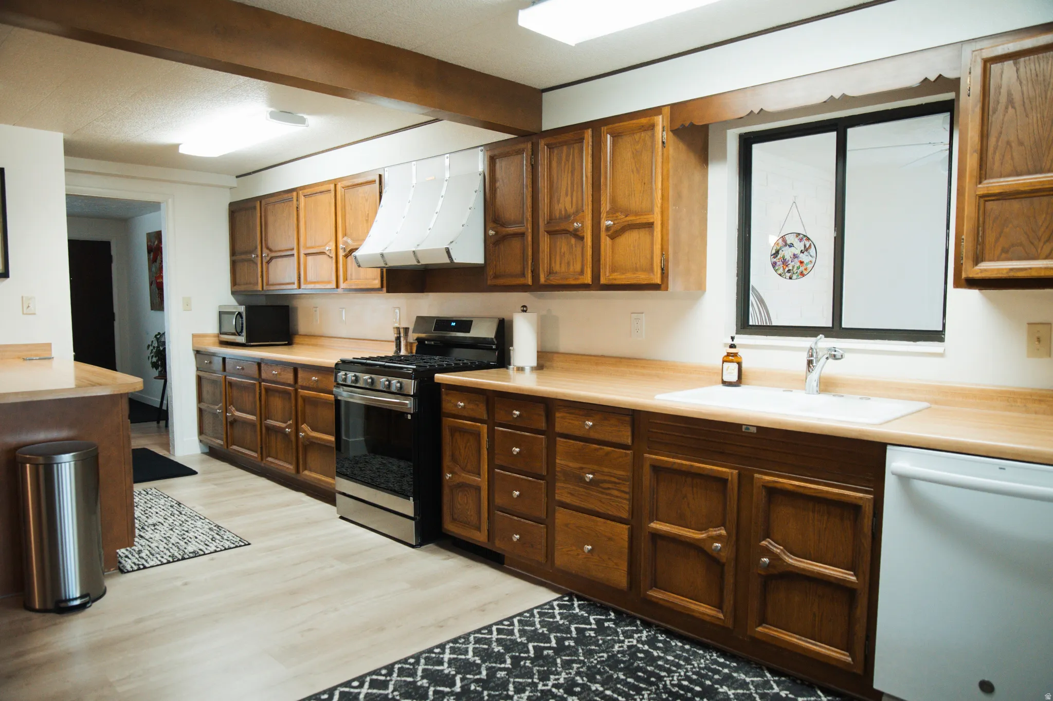 Kitchen featuring stainless steel appliances, light countertops, range hood, LVP floors, and beamed ceiling