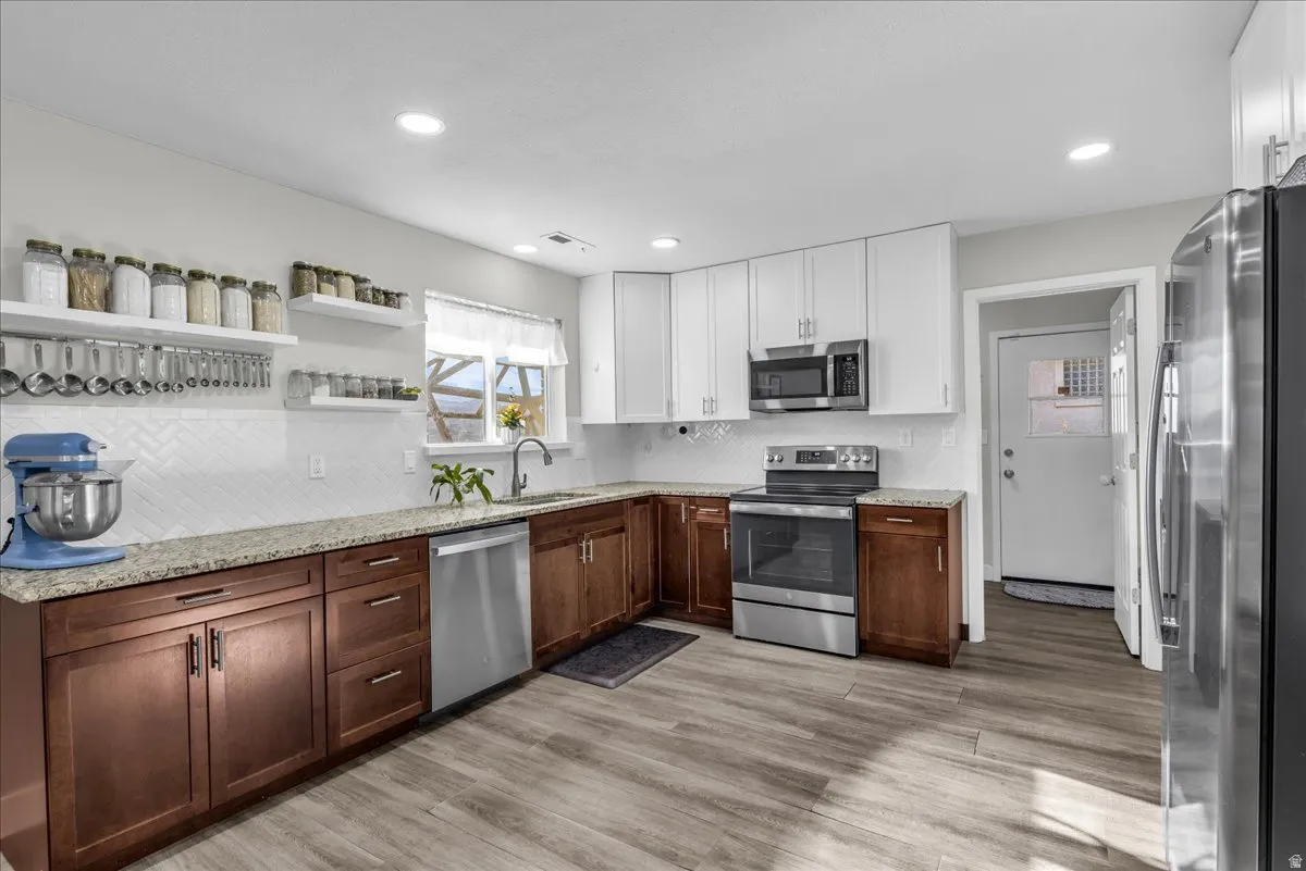 Two tone kitchen featuring open shelves, stainless steel appliances, two tone cabinets, light stone countertops, and decorative backsplash