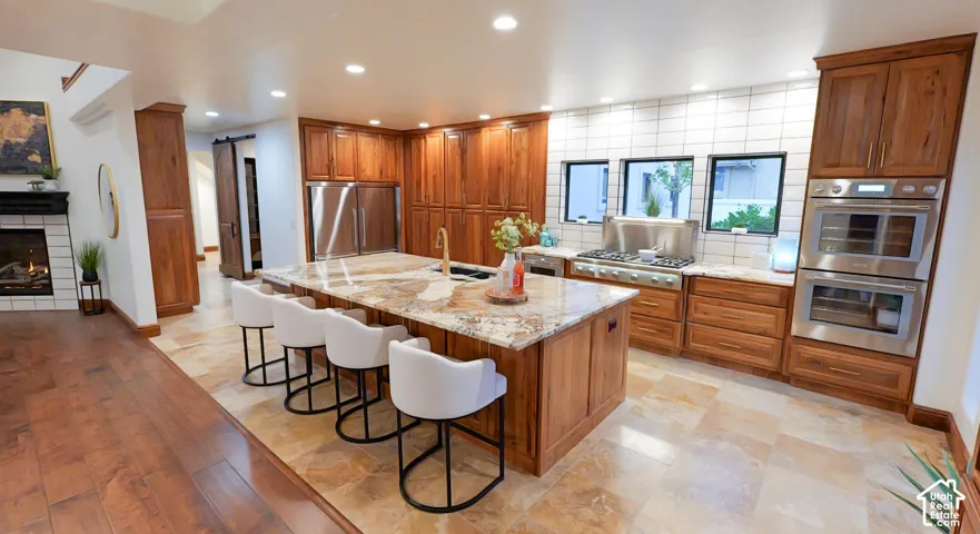 Kitchen featuring wood finish cabinets, light stone counters, stainless steel appliances, a kitchen breakfast bar, and recessed lighting