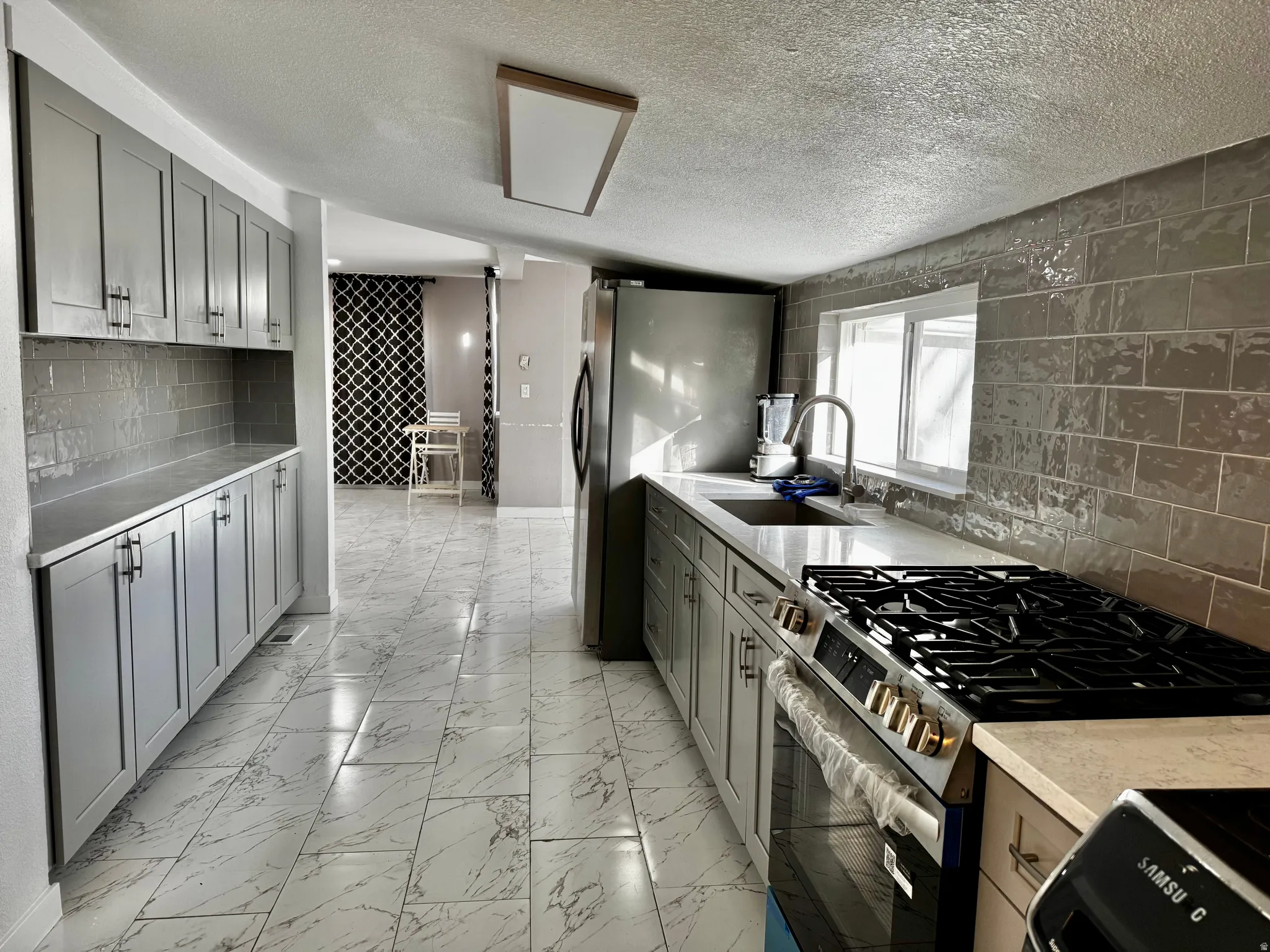 Kitchen featuring stainless steel appliances, gray cabinetry, backsplash, light marble finish flooring, and a textured ceiling